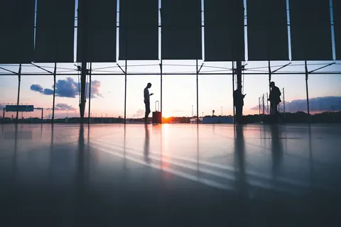 Travellers standing at the windows of an airport terminal with the sun low on the horizon